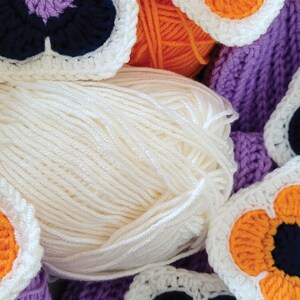 May include: Close-up of a ball of white yarn with crocheted flower motifs in purple, orange, and white. The flowers have black centers.