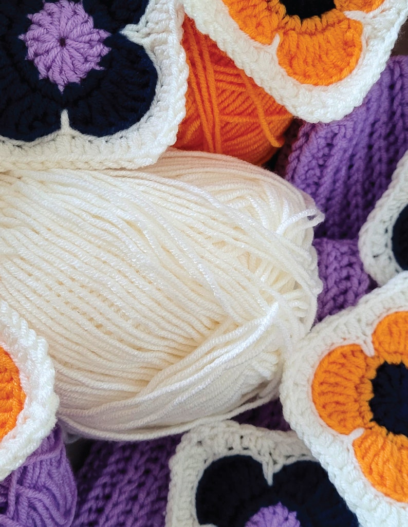 May include: Close-up of a ball of white yarn with crocheted flower motifs in purple, orange, and white. The flowers have black centers.