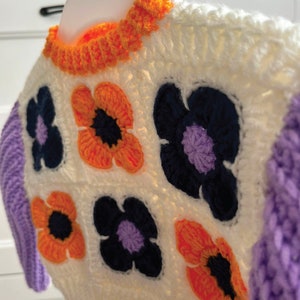 May include: A white crocheted sweater with a purple and orange floral pattern. The sweater has a white crocheted collar and long sleeves.