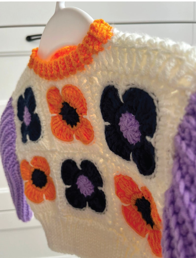 May include: A white crocheted sweater with a purple and orange floral pattern. The sweater has a white crocheted collar and long sleeves.