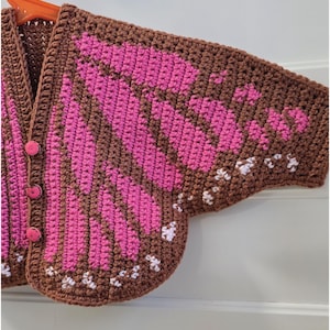 May include: A brown crochet cardigan featuring a vibrant pink butterfly wing pattern across the front and sleeves. The garment includes three round pink buttons along the front closure and small white stitched accents along the bottom edge.