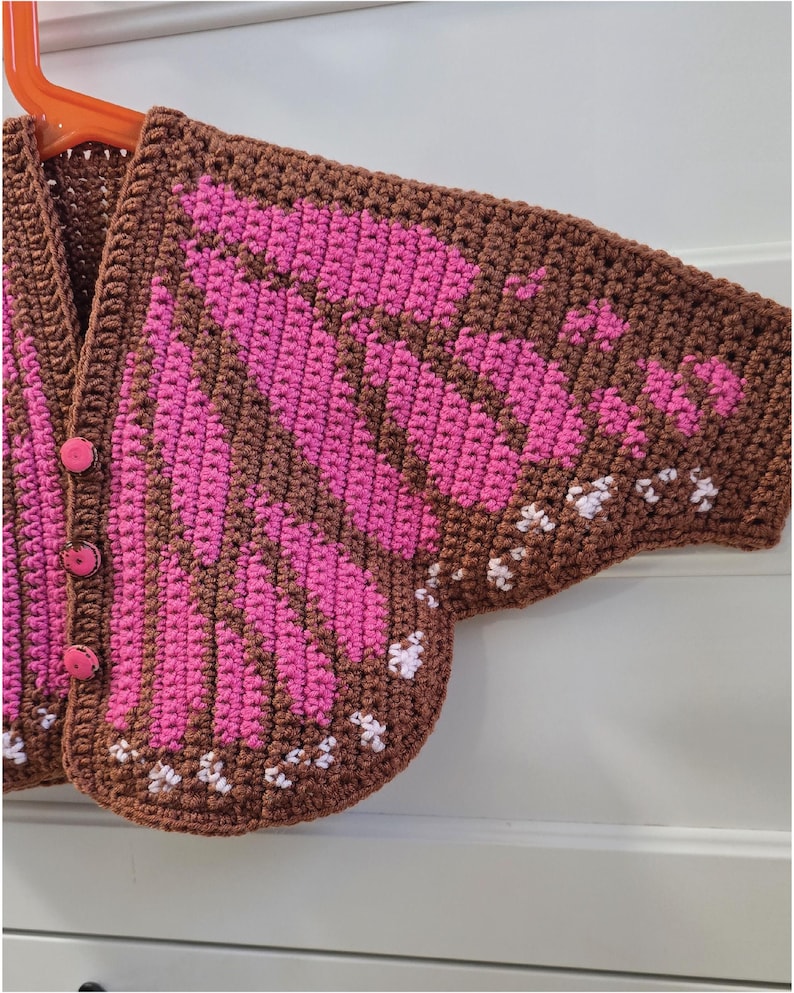 May include: A brown crochet cardigan featuring a vibrant pink butterfly wing pattern across the front and sleeves. The garment includes three round pink buttons along the front closure and small white stitched accents along the bottom edge.