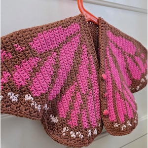 May include: A crochet cardigan featuring a brown base with vibrant pink wing-like patterns and small white accents along the bottom edges. The garment includes three matching pink buttons down the front and is displayed on an orange plastic hanger.