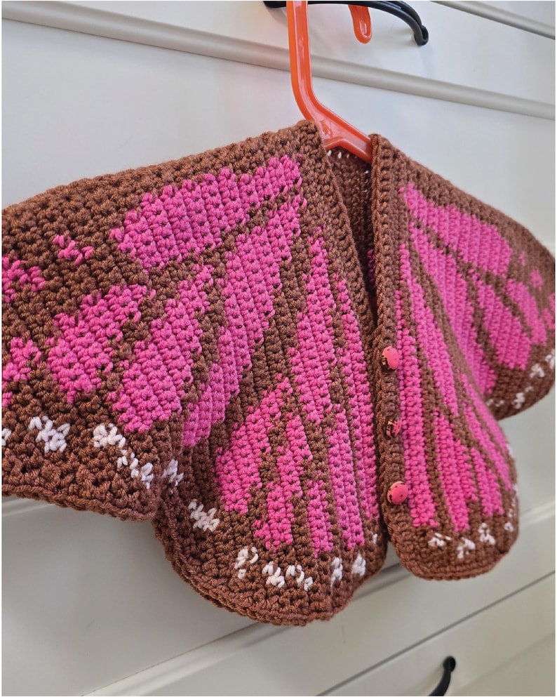 May include: A crochet cardigan featuring a brown base with vibrant pink wing-like patterns and small white accents along the bottom edges. The garment includes three matching pink buttons down the front and is displayed on an orange plastic hanger.