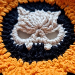 Peut inclure: Gros plan sur un motif de cr&acirc;ne crochet&eacute;. Le cr&acirc;ne est de couleur cr&egrave;me avec des accents noirs et orange. Le motif est circulaire, avec le cr&acirc;ne au centre, entour&eacute; d'un anneau noir et d'une bordure orange festonn&eacute;e.
