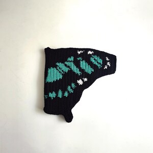 May include: A black, crocheted butterfly wing with teal and white accents. The wing features a detailed pattern of teal shapes and white floral-like designs, creating a textured appearance. The wing is set against a plain white background.