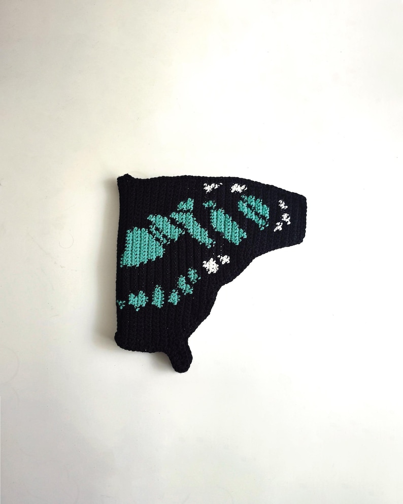 May include: A black, crocheted butterfly wing with teal and white accents. The wing features a detailed pattern of teal shapes and white floral-like designs, creating a textured appearance. The wing is set against a plain white background.