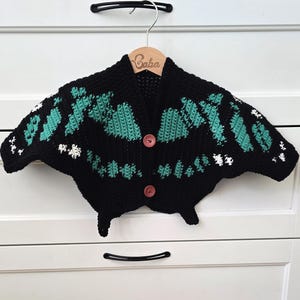 May include: A black crochet cardigan with a unique butterfly design, featuring teal and white accents. The cardigan has two pink buttons and is displayed on a wooden hanger with the name "Baba" engraved on it.