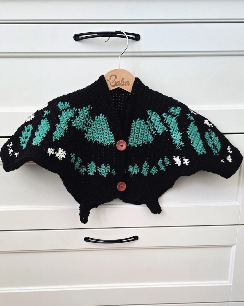 May include: A black crochet cardigan with a unique butterfly design, featuring teal and white accents. The cardigan has two pink buttons and is displayed on a wooden hanger with the name "Baba" engraved on it.