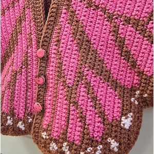 May include: A crochet cardigan featuring a vibrant butterfly wing pattern in shades of brown and bright pink. The garment includes three round pink buttons down the front placket and small white stitched accents along the bottom edge of the wings.