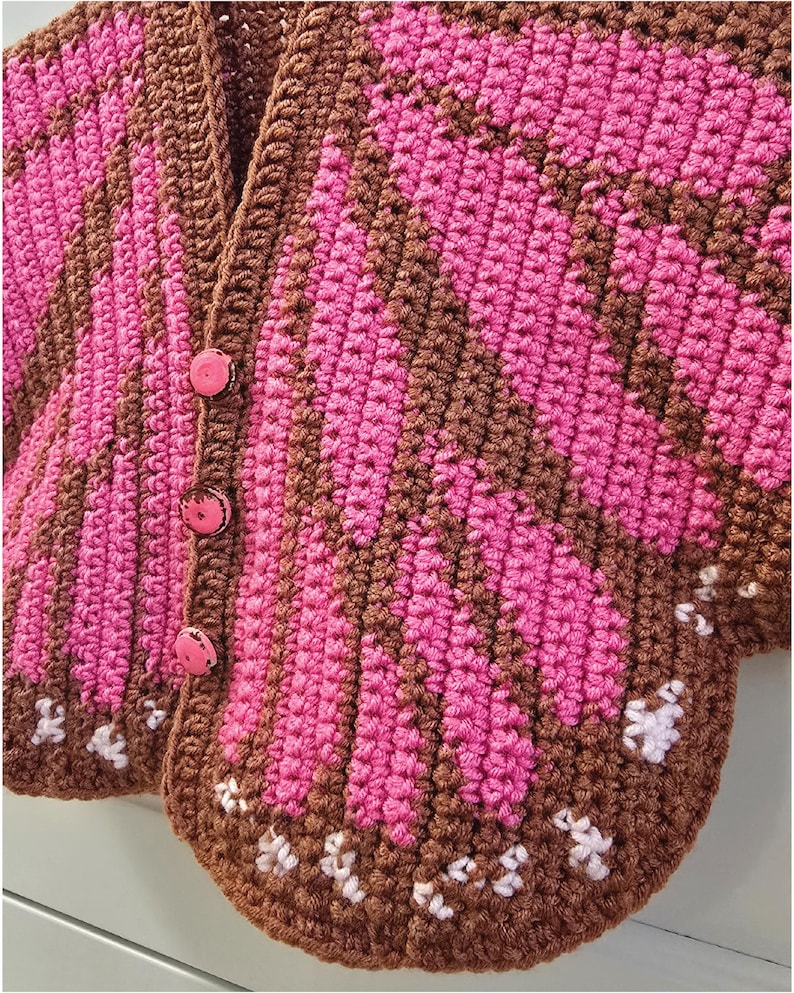 May include: A crochet cardigan featuring a vibrant butterfly wing pattern in shades of brown and bright pink. The garment includes three round pink buttons down the front placket and small white stitched accents along the bottom edge of the wings.