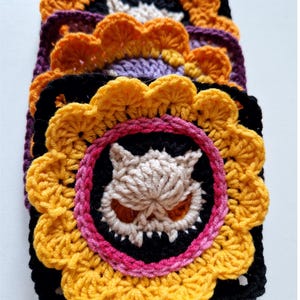 Peut inclure: Une pile de carr&eacute;s crochet&eacute;s faits &agrave; la main de diff&eacute;rentes couleurs. Chaque carr&eacute; pr&eacute;sente un motif central de cr&acirc;ne aux yeux oranges et aux dents pointues, entour&eacute; d'un cercle rose et noir, et d'une bordure jaune en forme de fleur. Les carr&eacute;s sont empil&eacute;s.