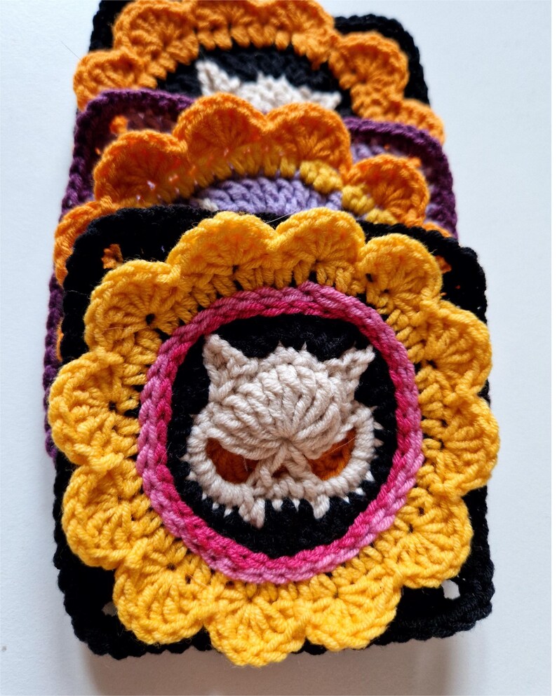 Peut inclure: Une pile de carr&eacute;s crochet&eacute;s faits &agrave; la main de diff&eacute;rentes couleurs. Chaque carr&eacute; pr&eacute;sente un motif central de cr&acirc;ne aux yeux oranges et aux dents pointues, entour&eacute; d'un cercle rose et noir, et d'une bordure jaune en forme de fleur. Les carr&eacute;s sont empil&eacute;s.