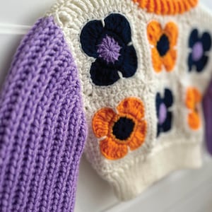 May include: A white crocheted sweater with purple, orange, and blue flower accents. The sweater has long sleeves and a crew neck.