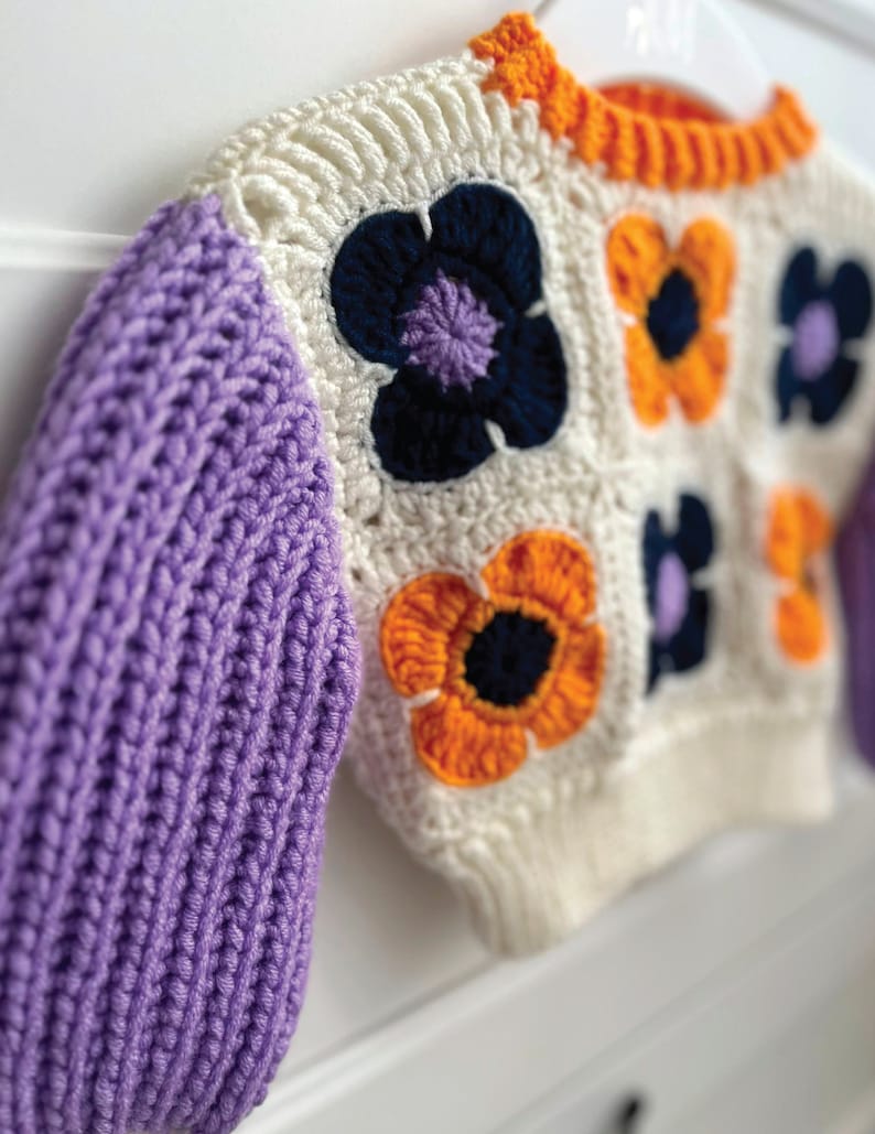 May include: A white crocheted sweater with purple, orange, and blue flower accents. The sweater has long sleeves and a crew neck.