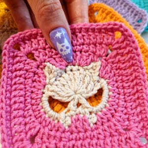 May include: A close-up of a pink crocheted square with a skull design in the center. The skull is made of off-white yarn and has orange accents. Other colorful crocheted squares are visible in the background.