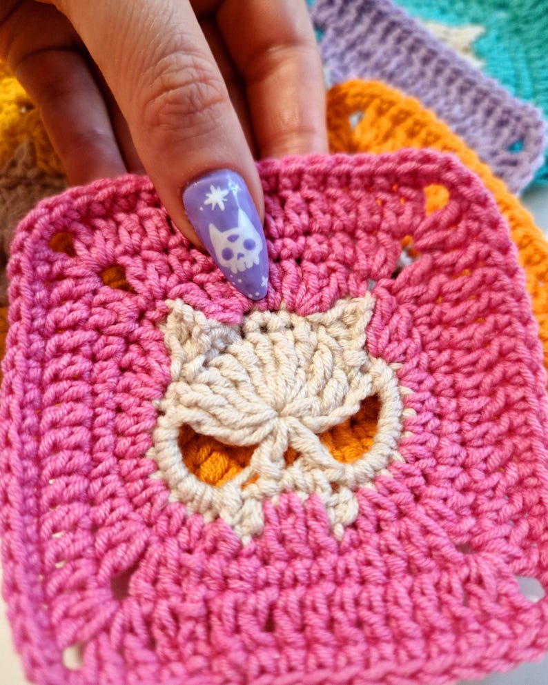 May include: A close-up of a pink crocheted square with a skull design in the center. The skull is made of off-white yarn and has orange accents. Other colorful crocheted squares are visible in the background.