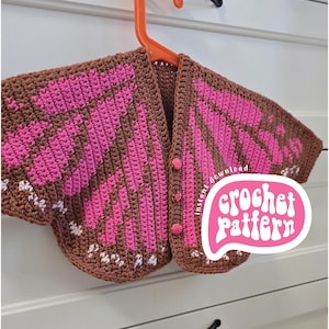Op de afbeelding: Een bruin en roze gehaakt vest met een vlindervleugelontwerp, hangend aan een oranje hanger. Het vest heeft drie roze knopen en witte bloemaccenten. De tekst "crochet pattern" is zichtbaar.
