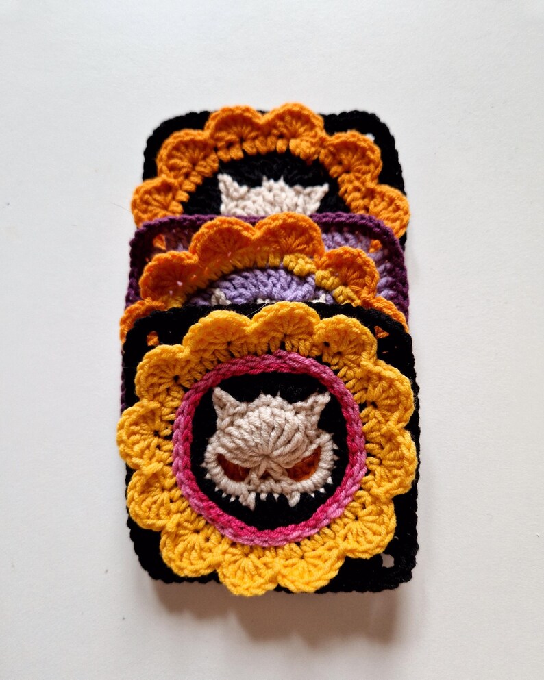 Peut inclure: Une pile de quatre dessous de verre crochet&eacute;s. Chaque dessous de verre est carr&eacute; avec une bordure noire et un motif central repr&eacute;sentant un cr&acirc;ne de dessin anim&eacute;. Le cr&acirc;ne est entour&eacute; d'un motif floral jaune et orange. Les dessous de verre sont empil&eacute;s sur une surface blanche.
