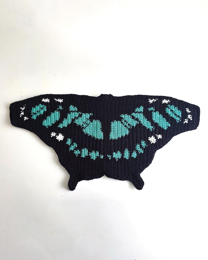 May include: A black, butterfly-shaped crochet piece with turquoise and white accents. The wings feature a detailed pattern, and the overall design is symmetrical. The piece is likely handmade and suitable for decorative purposes.