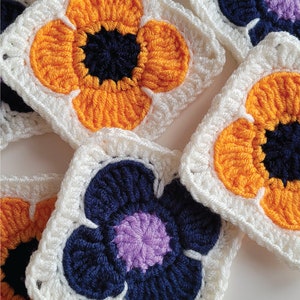 May include: Crocheted flower squares in white, orange, purple, and blue. The squares are made with a granny stitch pattern and feature a flower design in the center.