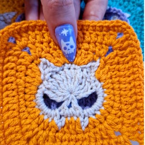May include: A crocheted square in bright orange with a skull design in the center. The skull is off-white with dark purple eye sockets. The square is held up by a hand with a blue nail with a skull design.