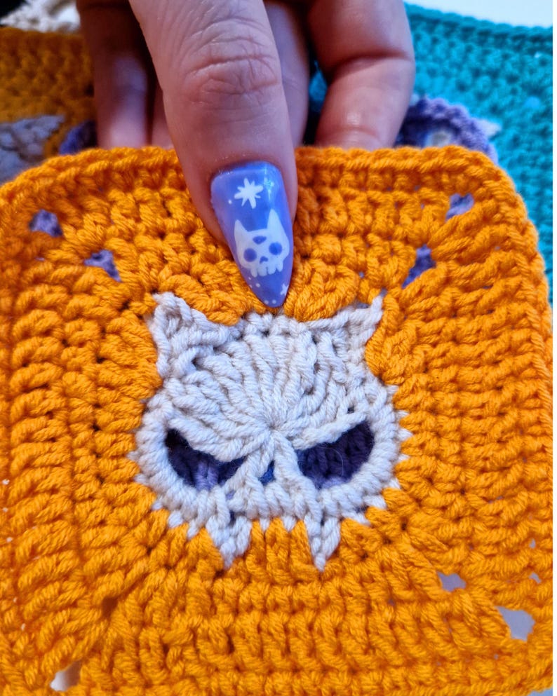 May include: A crocheted square in bright orange with a skull design in the center. The skull is off-white with dark purple eye sockets. The square is held up by a hand with a blue nail with a skull design.
