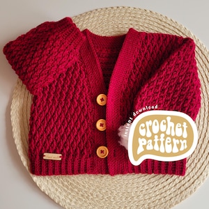 May include: A red crochet cardigan with a textured diamond pattern. The cardigan has three wooden buttons and a small wooden tag that says "Hand made". A sticker that says "crochet pattern" is on the right side of the cardigan.