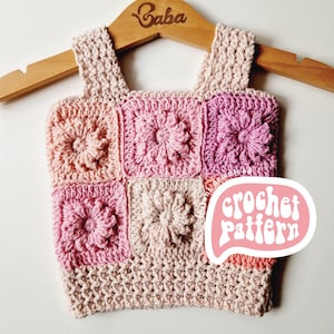 Könnte beinhalten: Ein Häkeltop mit einem quadratischen Muster in Rosatönen und Pfirsich. Das Oberteil hat ein gehäkeltes Blumendesign und hängt an einem Holzbügel mit dem eingravierten Wort "Baba". Der Text "crochet pattern" ist in einer Sprechblase geschrieben.