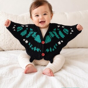 May include: A smiling baby wearing a black knitted cardigan with a butterfly design. The cardigan features teal and white accents and red buttons. The baby is also wearing white pants.