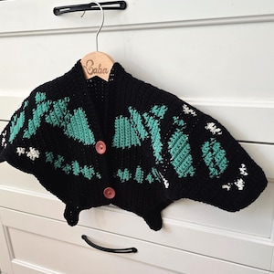 May include: A black crochet cardigan with teal and white accents, featuring a unique butterfly-like design. The cardigan has two brown buttons and is displayed on a wooden hanger with the name "Gaba" engraved on it.