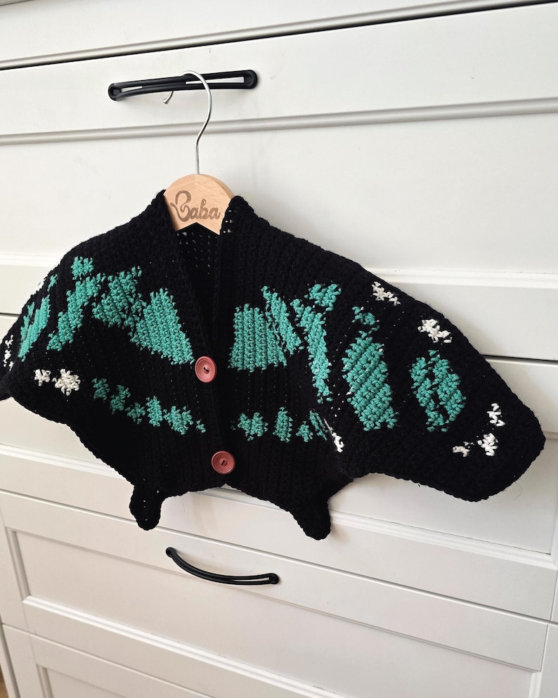 May include: A black crochet cardigan with teal and white accents, featuring a unique butterfly-like design. The cardigan has two brown buttons and is displayed on a wooden hanger with the name "Gaba" engraved on it.