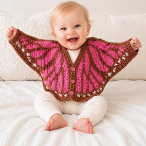 May include: A crochet cardigan featuring a butterfly wing pattern in shades of brown and bright pink. The garment has long sleeves, a V-neckline, and three small pink buttons down the front. The edges are detailed with small white accents.