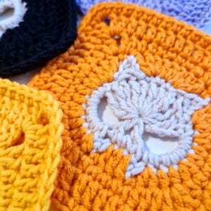 May include: Close-up of several crocheted squares in various colors, including orange, yellow, black, and purple. Each square features a white owl design in the center. The handmade squares are likely part of a larger craft project.