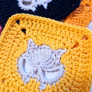 May include: Close-up of several crocheted squares with a cat face design. The squares are in various colors, including yellow, black, and orange. The cat faces are in a contrasting off-white color, creating a striking visual contrast. The crochet work appears to be handmade.