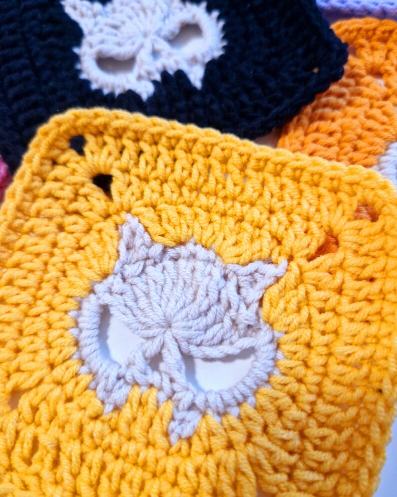 May include: Close-up of several crocheted squares with a cat face design. The squares are in various colors, including yellow, black, and orange. The cat faces are in a contrasting off-white color, creating a striking visual contrast. The crochet work appears to be handmade.