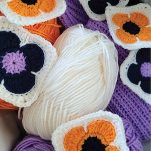 May include: A close-up of a collection of crocheted flower motifs in shades of orange, purple, and white. The flowers are arranged around balls of yarn in purple and white.