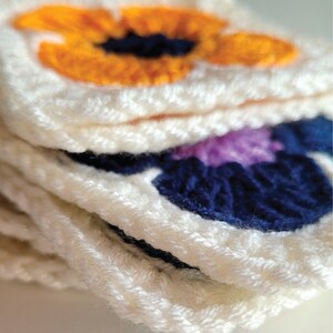 May include: A stack of crocheted squares with floral designs. The top square features a bright orange flower with a dark blue center. The next square features a dark blue flower with a purple center.