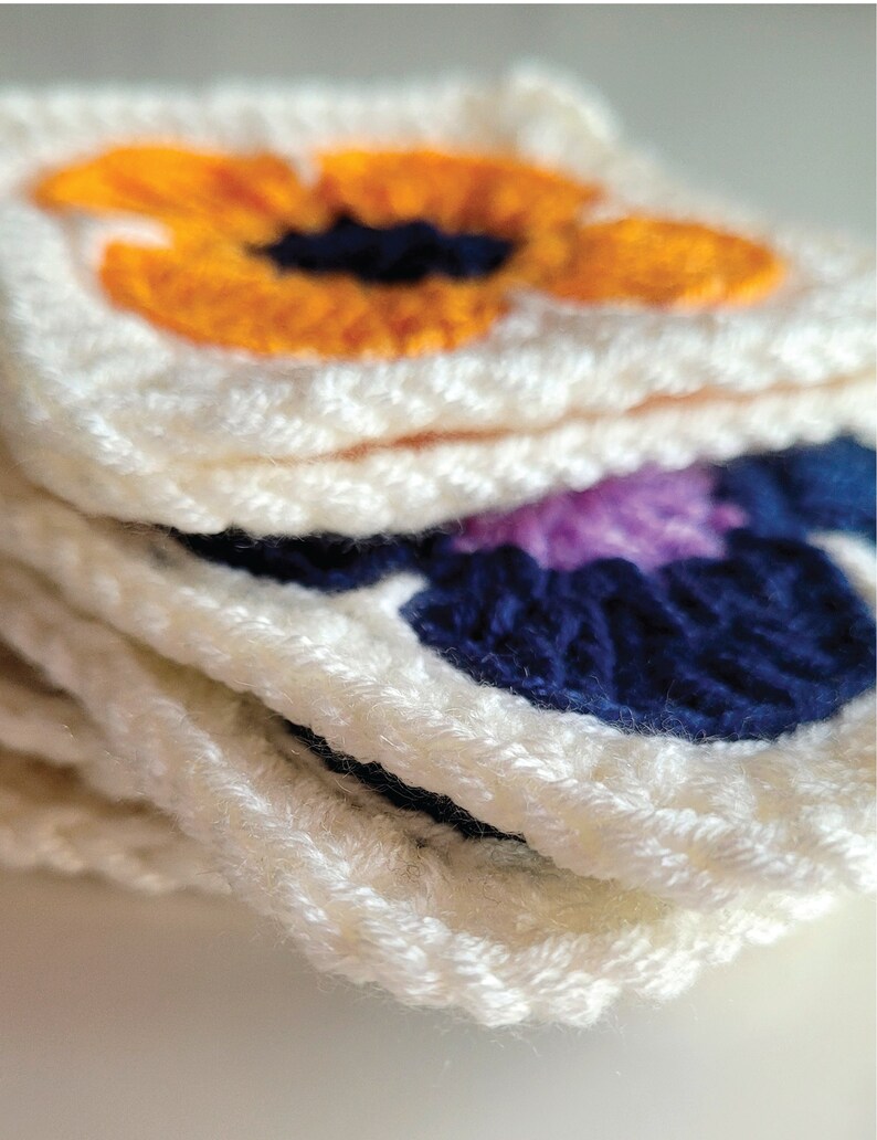 May include: A stack of crocheted squares with floral designs. The top square features a bright orange flower with a dark blue center. The next square features a dark blue flower with a purple center.