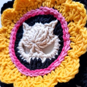 Peut inclure: Une fleur au crochet faite &agrave; la main avec un motif de cr&acirc;ne central. La fleur pr&eacute;sente un sch&eacute;ma de couleurs jaune, noir, rose et cr&egrave;me. Le cr&acirc;ne est de couleur cr&egrave;me, entour&eacute; d'un cercle noir et d'une bordure rose. Les p&eacute;tales ext&eacute;rieurs sont jaunes.