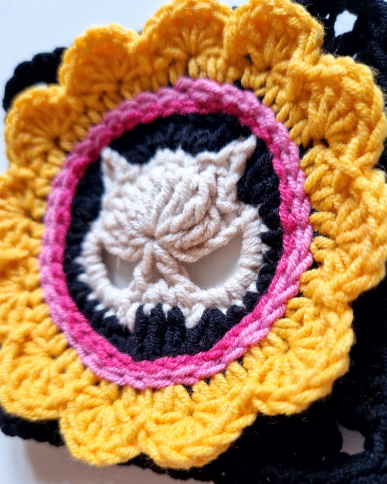 Peut inclure: Une fleur au crochet faite &agrave; la main avec un motif de cr&acirc;ne central. La fleur pr&eacute;sente un sch&eacute;ma de couleurs jaune, noir, rose et cr&egrave;me. Le cr&acirc;ne est de couleur cr&egrave;me, entour&eacute; d'un cercle noir et d'une bordure rose. Les p&eacute;tales ext&eacute;rieurs sont jaunes.