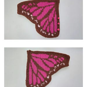 May include: A pair of crocheted butterfly wings in brown and bright pink. The wings feature a dark brown border with a pink interior pattern and small white accents along the outer edge. Three small pink buttons are attached to the side edge.