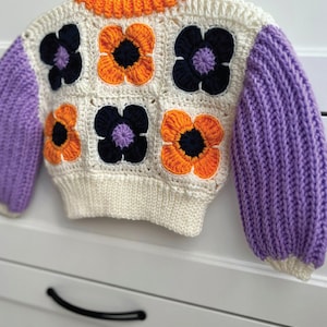 May include: A white crocheted sweater with orange, purple, and black flower accents. The sweater has long sleeves and a round neckline.