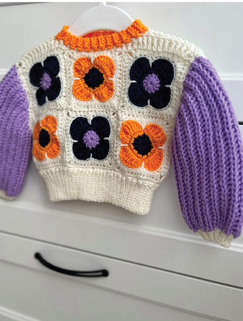 May include: A white crocheted sweater with orange, purple, and black flower accents. The sweater has long sleeves and a round neckline.