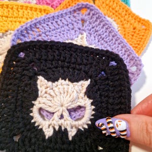 May include: A stack of colorful crochet squares, each featuring a skull design. The squares are in various colors, including black, purple, orange, and pink. The skull is a contrasting color, adding detail to the handmade craft.