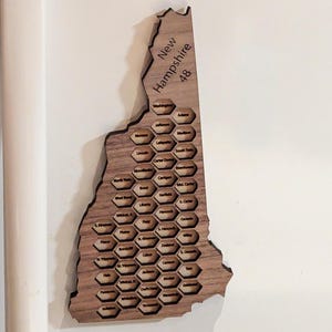 May include: A wooden map of New Hampshire with the state's counties labeled in a honeycomb pattern. The map is made of light brown wood and has a natural wood grain.