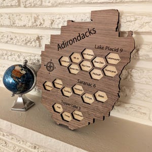 May include: A wooden map of the Adirondacks region with engraved place names and a compass rose. The map includes the text "Adirondacks", "Lake Placid 9", "Saranac 6", and "Tupper Lake 3". A small globe sits to the left.