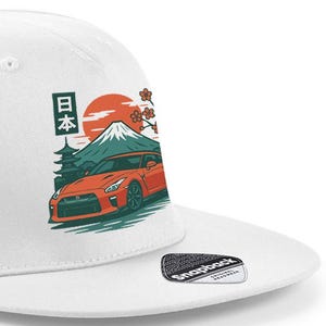Peut inclure: Casquette de baseball blanche avec un motif d'une voiture de sport rouge, du Mont Fuji et du texte japonais. Le design comprend un soleil levant et des fleurs de cerisier. La casquette a une visière plate et une fermeture à pression.