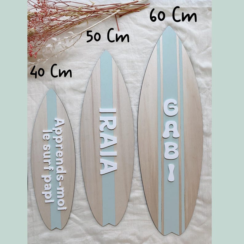 以下が含まれることがあります： 木製のサーフボード3枚。長さは40cm、50cm、60cmで、水色のストライプと白い文字が特徴です。一番小さいボードには「Apprends-moi le surf papi」と書かれています。真ん中のボードには「IRAIA」、一番大きいボードには「GABI」と書かれています。