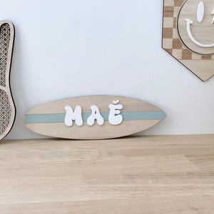 以下が含まれることがあります： 白いバブルスタイルの文字で「MAE」という名前が書かれた、サーフボード型の木製サイン。薄い青色のストライプがサインの中央を横切っています。平和のサインやスマイリーなど、他の木製の装飾品が背景にあります。